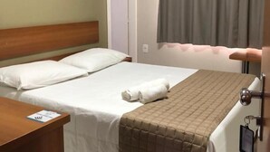 Una habitación de hotel con cama, mesita de noche, equipo de aire acondicionado, ventana con cortinas y puerta.