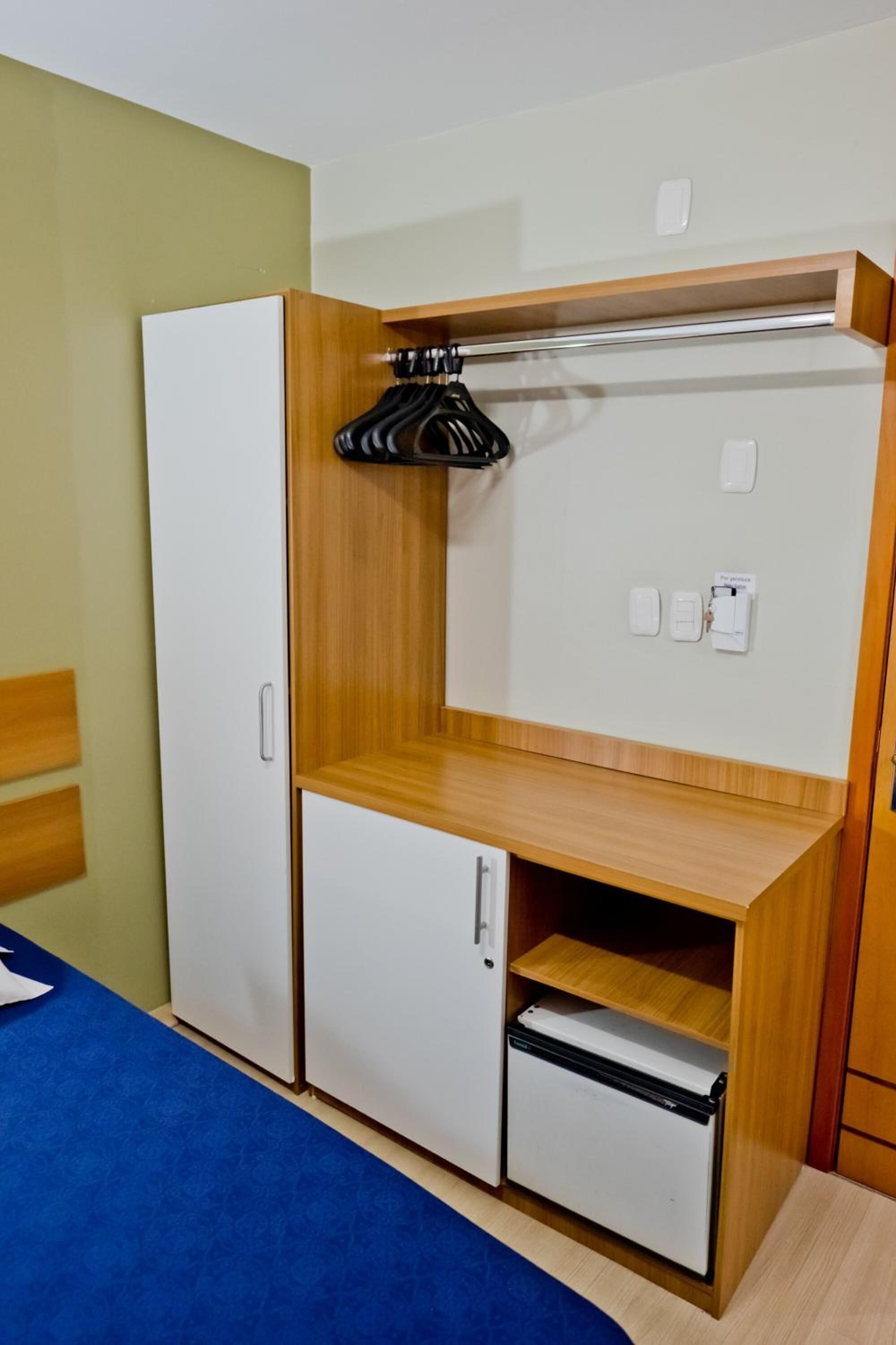 A compact bedroom with a bed, a wardrobe, a small desk, and a shelf.