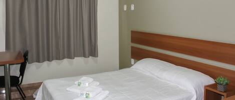 A neatly made bed with white linens and towels, a wooden headboard, a small bedside table with a plant, and a chair in a simple room with a curtain and an air conditioning unit.