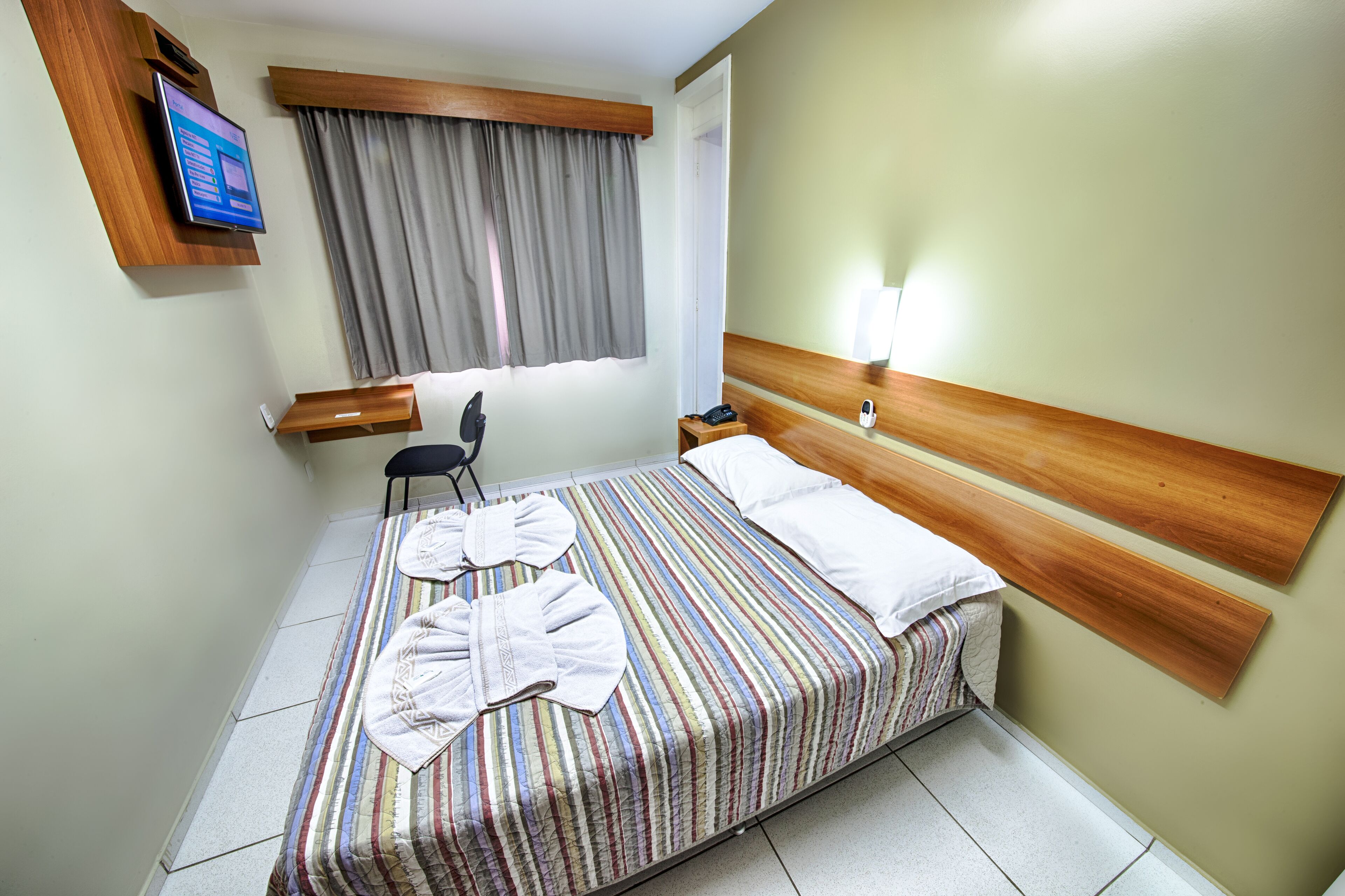 A hotel room with a bed, two pillows, a TV, a chair, and a window with curtains.