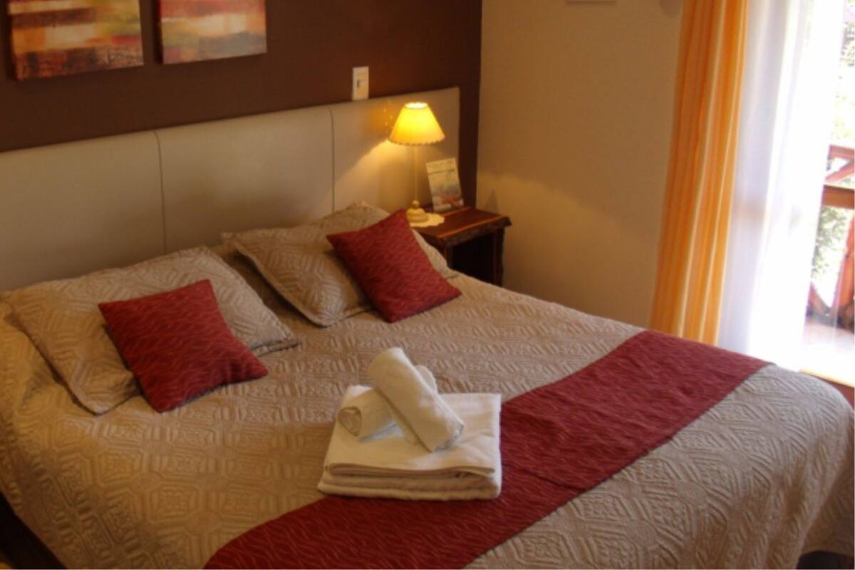 A neatly made bed with a red and beige bedspread, two red pillows, and a folded towel on the bed. There are two bedside tables with lamps and a phone on one of them.
