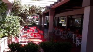 An outdoor seating area with red tables and chairs, some with Coca-Cola branding, under a covered structure with a view of the street.