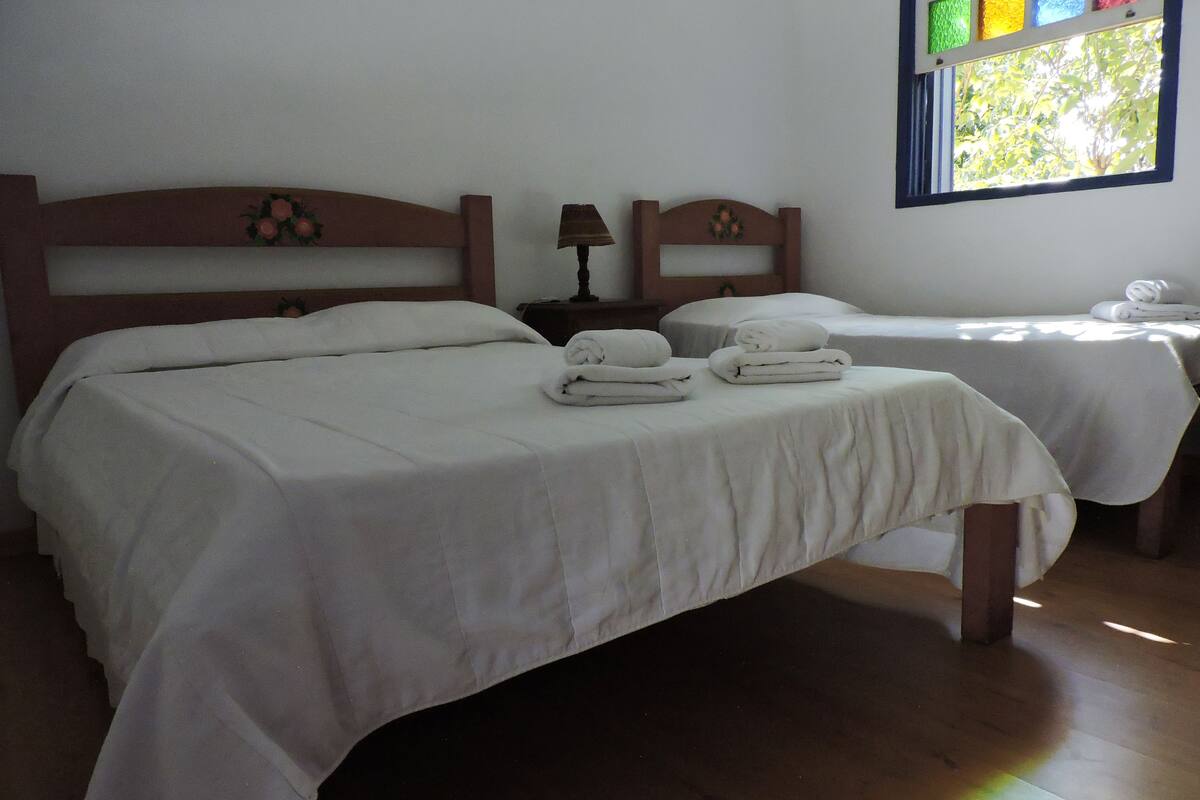 A hotel room with two beds, each with white linens and pillows, a small table with a lamp, and a window with a view of greenery.