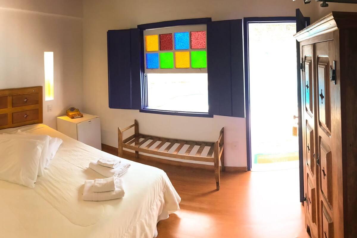 A bedroom with a bed, bedside table, lamp, wardrobe, and a window with colorful panels.