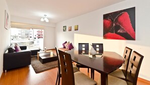 A modern living room with a dining area, featuring a large window, a black sofa, a wooden dining table, and a red abstract painting on the wall. - Wasi Apartments Atahualpa (Lima)