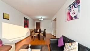 A modern living room with a black leather sofa, a dining table with chairs, and a colorful abstract painting on the wall. - Wasi Apartments Atahualpa (Lima)