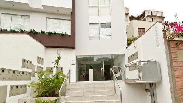 A modern building with a staircase leading to a glass entrance, surrounded by greenery and a brick wall. - Wasi Apartments Atahualpa (Lima)
