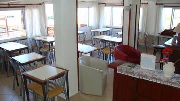 Un restaurante con varias mesas y sillas, una barra con un jarrón de flores y grandes ventanales con cortinas.
