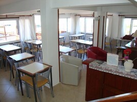Un restaurante con varias mesas y sillas, una barra con un jarrón de flores y grandes ventanales con cortinas.