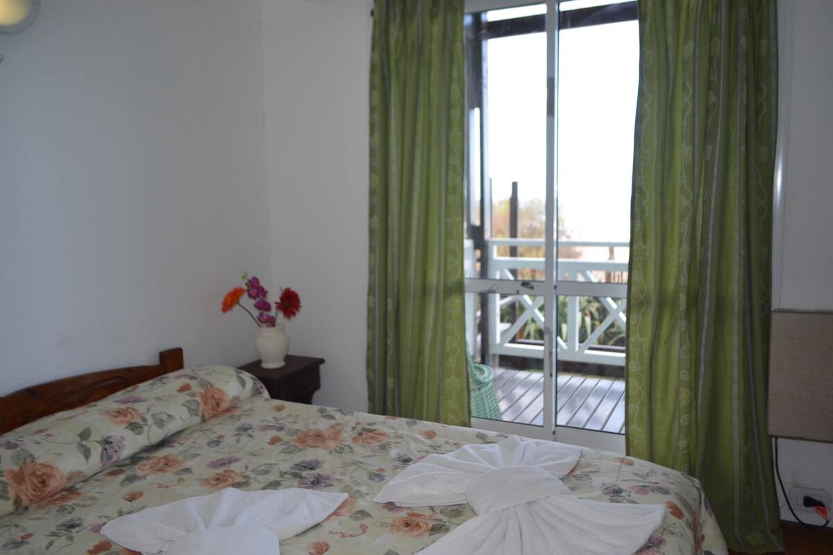 Un dormitorio con cama, colcha estampada, un florero con flores y un balcón con cortinas verdes.