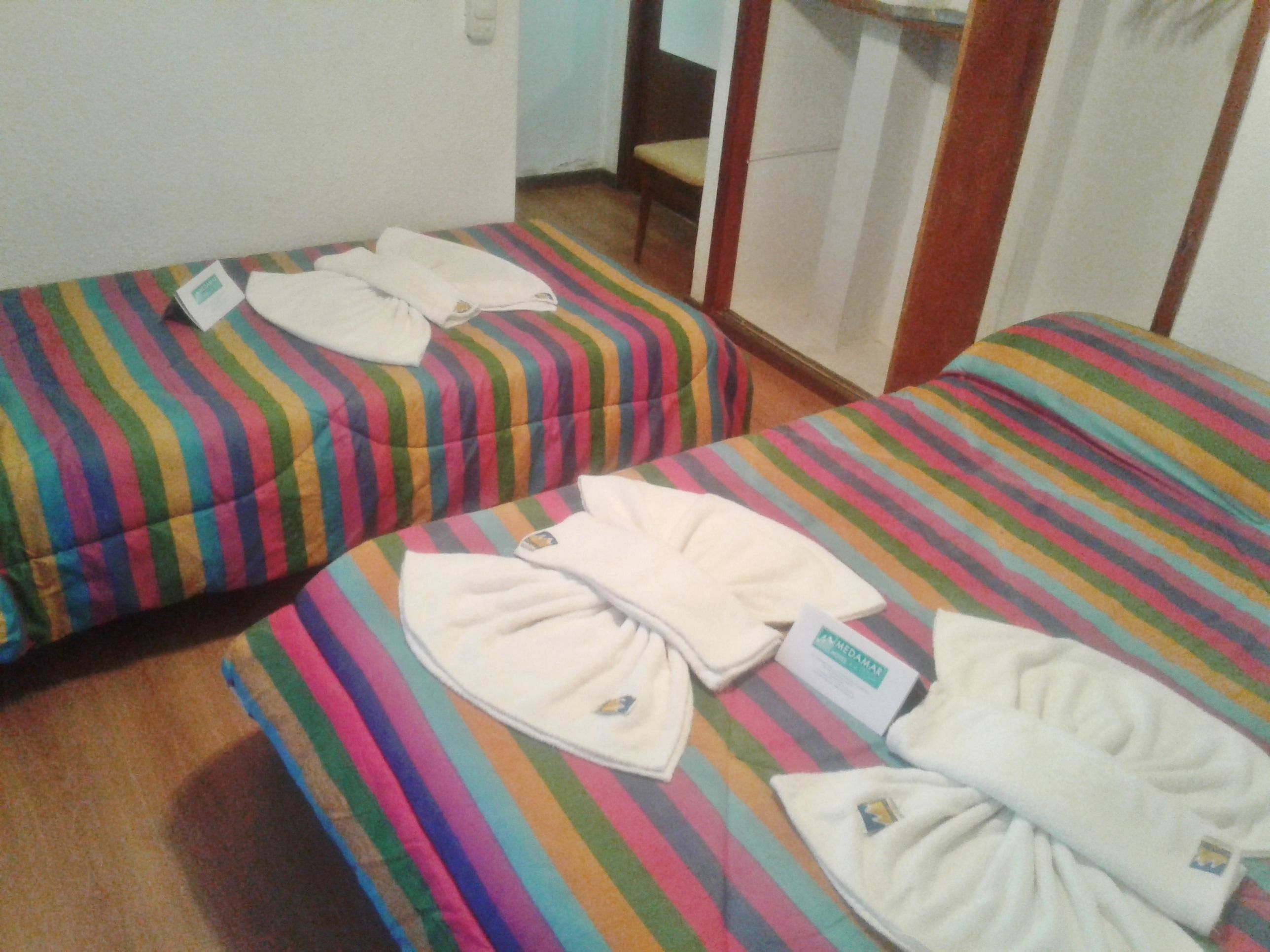 Duas camas com listras, almofadas e toalhas dobradas, uma porta de madeira e uma pequena mesa com uma cadeira.