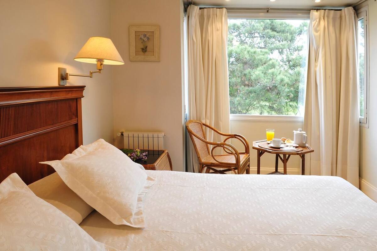 Quarto de hotel com cama, cadeira, mesa pequena com itens de café da manhã, luminária e janela com cortinas.