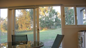 Um quarto com mesa e cadeiras de vidro, porta de correr de vidro que dá para uma área externa gramada, e uma cozinha com fogão e forno.
