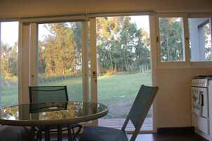 Una habitación con una mesa y sillas de vidrio, una puerta corrediza de vidrio que da a un área verde al aire libre, y una cocina con estufa y horno.