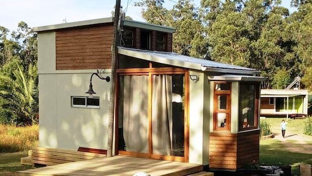 Uma cabana pequena, térrea, com um deck de madeira, porta de vidro deslizante e um pequeno alpendre.