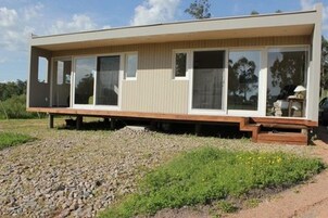 Una casa moderna de un piso con amplios ventanales y techo plano, ubicada en un camino de grava rodeado de vegetación.