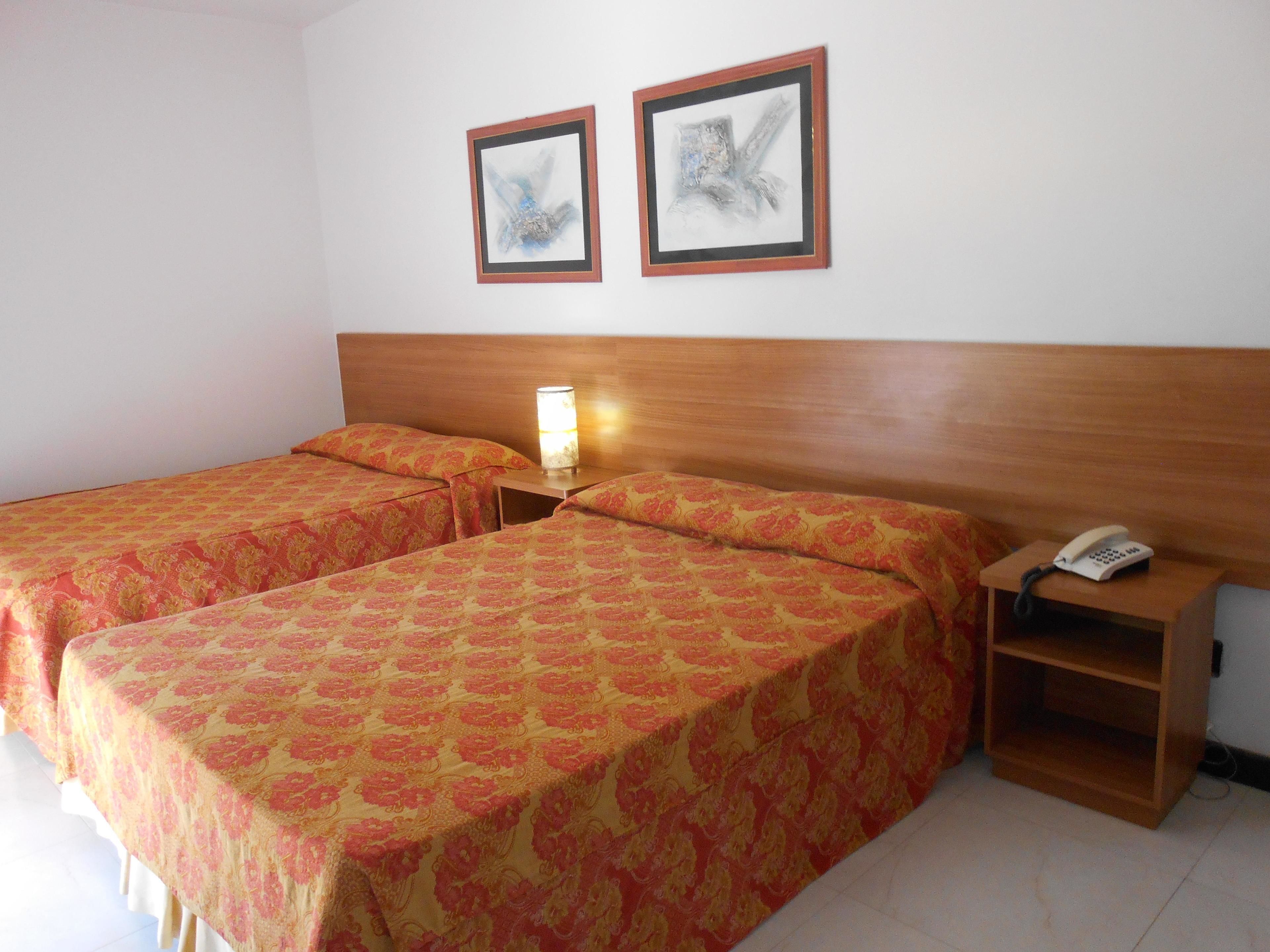 A hotel room with two single beds, a bedside table with a lamp, a phone, and two framed pictures on the wall.