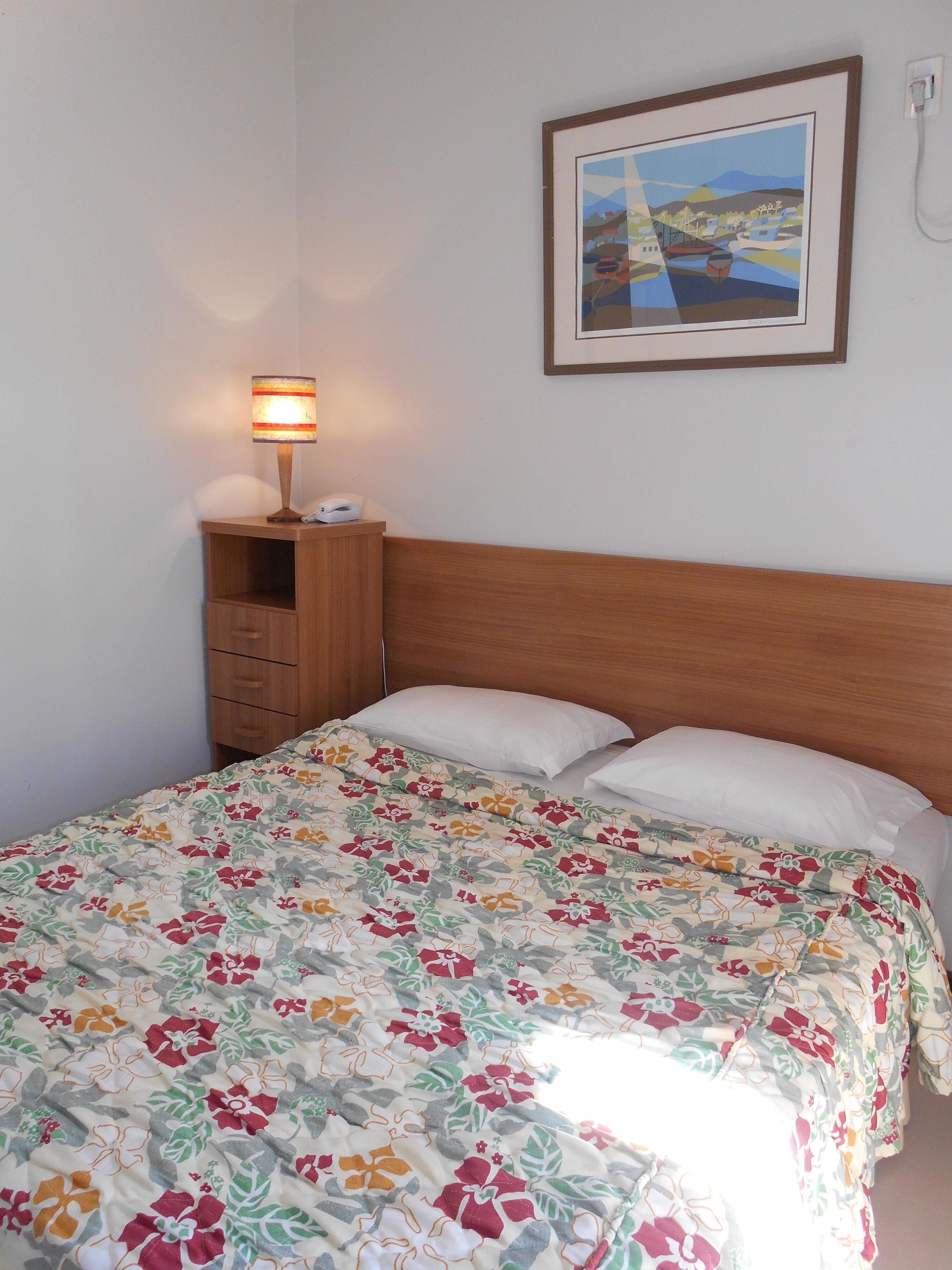 A neatly made bed with a floral bedspread, a bedside lamp, a small wooden nightstand with a phone, and a framed picture on the wall.