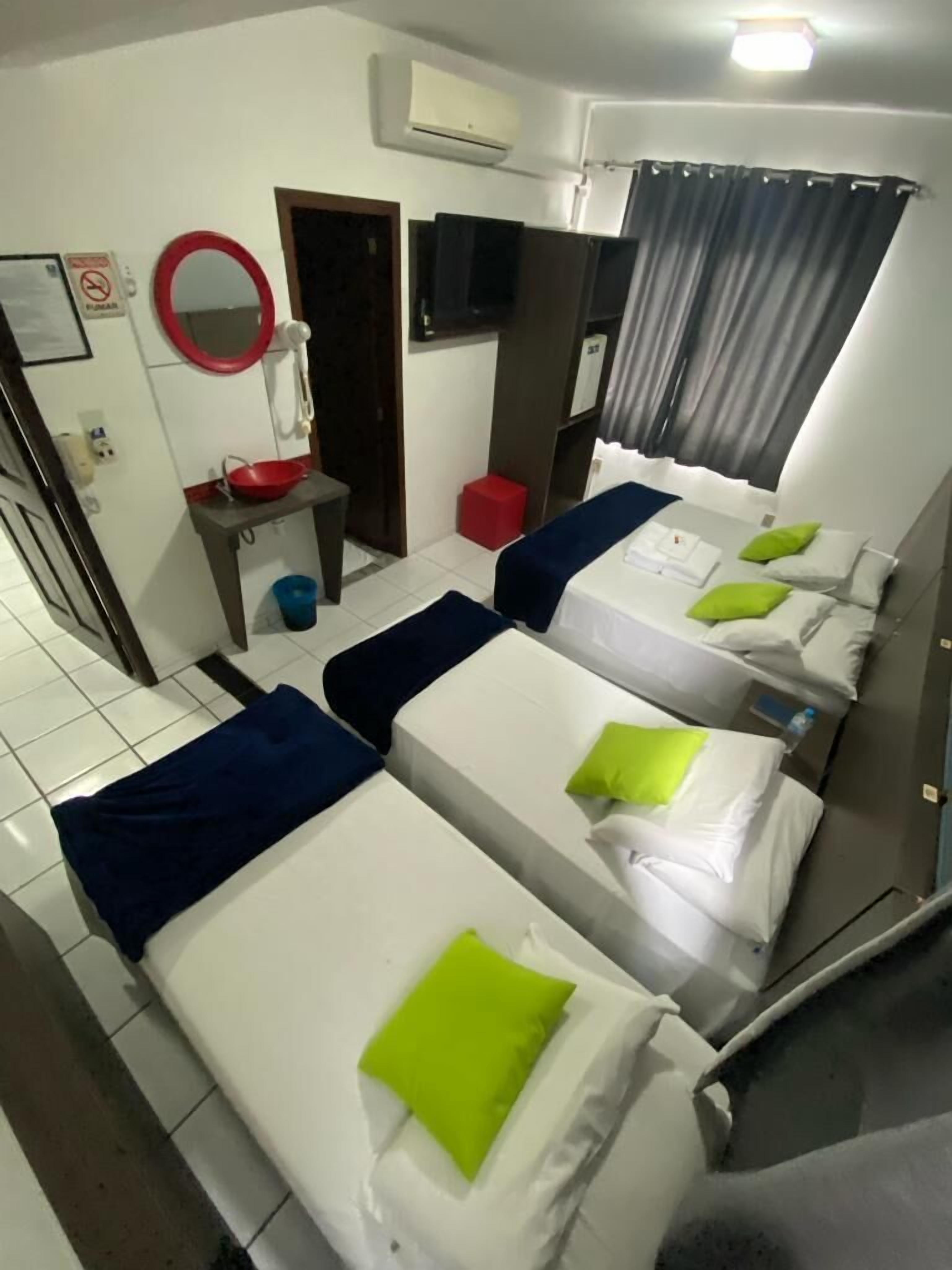 Habitación de hotel con cuatro camas individuales, una mesita con un cuenco rojo, un espejo, un equipo de aire acondicionado y una televisión.