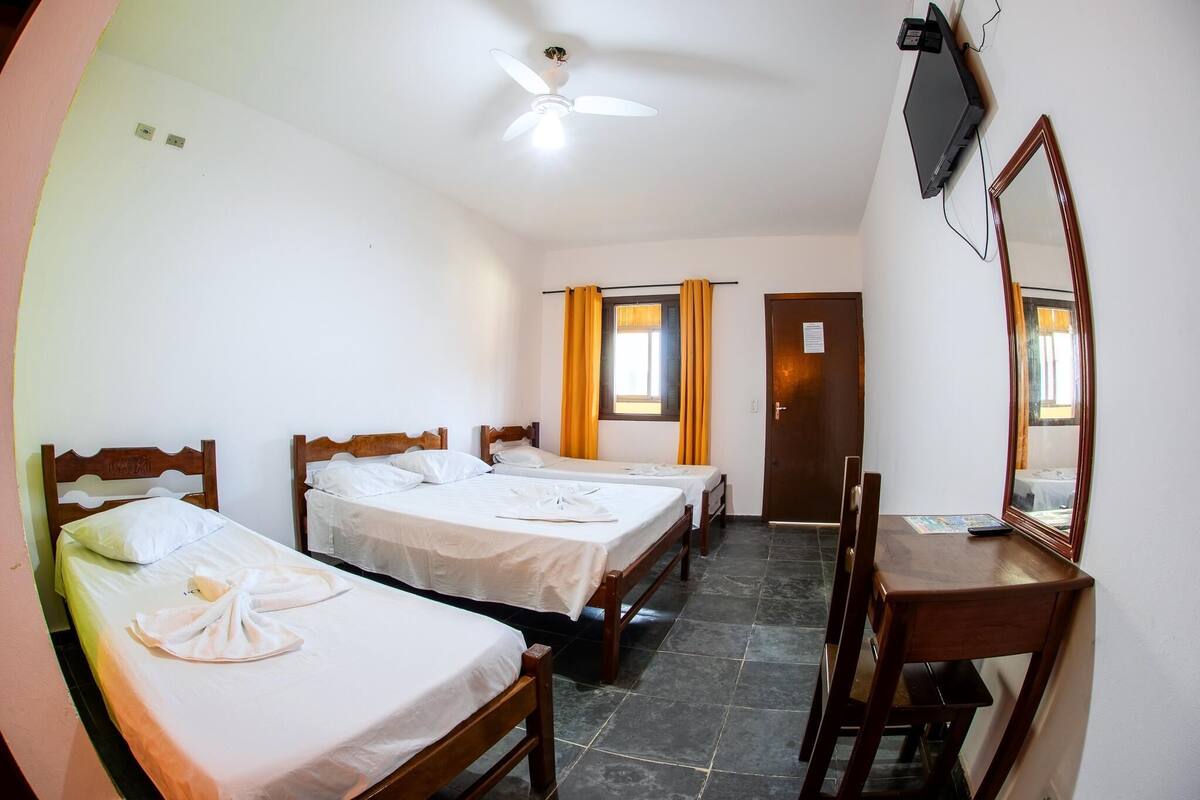 Une chambre d’hôtel avec deux lits simples, un bureau avec une chaise, un téléviseur fixé au mur et un ventilateur de plafond.