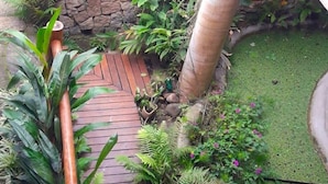 Una terraza de madera que conduce a un muro de piedra, rodeada de exuberante vegetación tropical y una pequeña zona con césped.
