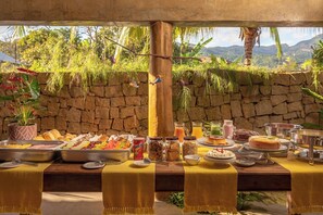 A buffet table with a variety of food items and drinks, set against a stone wall with a view of mountains and greenery.