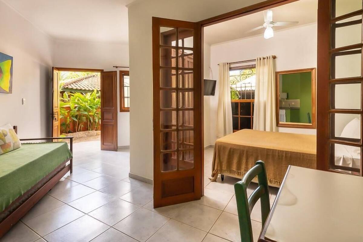 A room with a bed, a table, and a chair. There is a door leading to a balcony with plants.