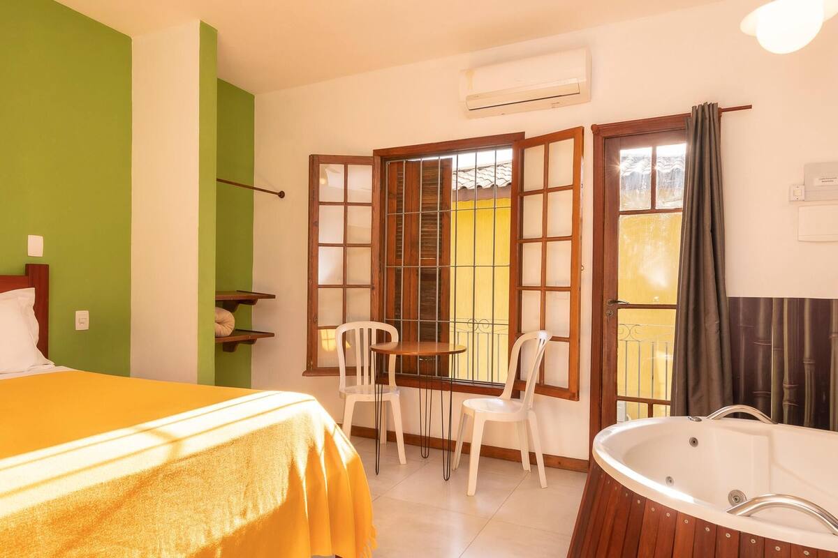 A room with a bed, a small table with chairs, a bathtub, and a window with a view of a yellow building.