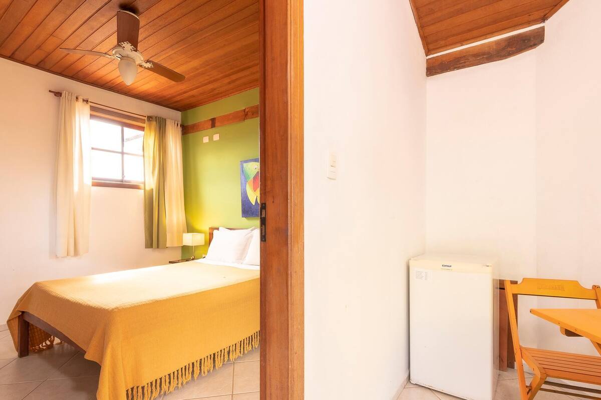 A room with a bed, a desk, a chair, a ceiling fan, and a refrigerator.
