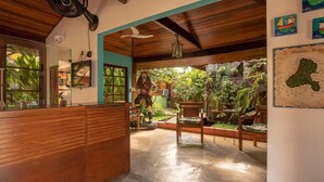 Zona de recepción con un mostrador de madera, ventilador de techo y vistas a un exuberante jardín con una estatua.