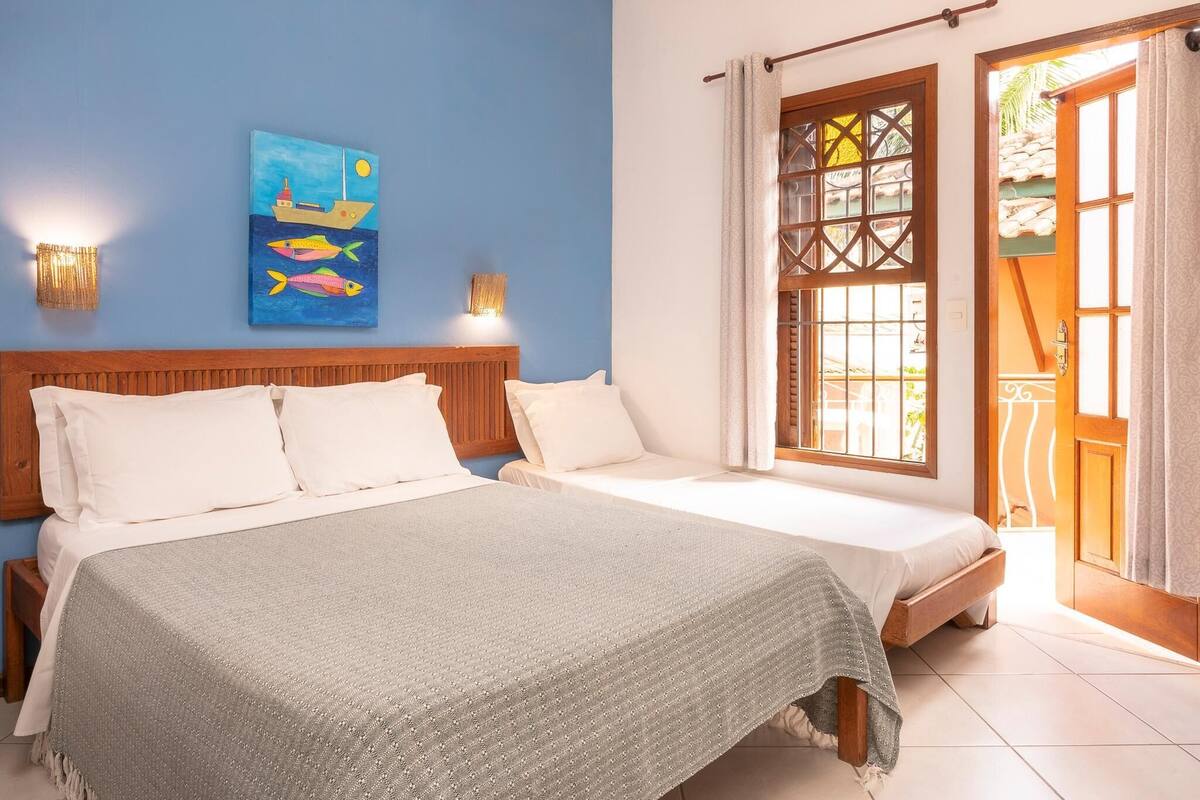 A bedroom with two beds, a blue wall, a painting of fish, and a window with curtains.
