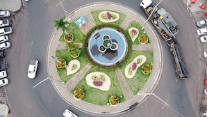 Une place circulaire avec une fontaine centrale, entourée de jardins aménagés et d’un espace de stationnement.