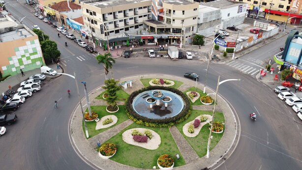 Una glorieta con una fuente central rodeada de macizos de flores, caminos que se cruzan y edificios circundantes, incluyendo un hotel.