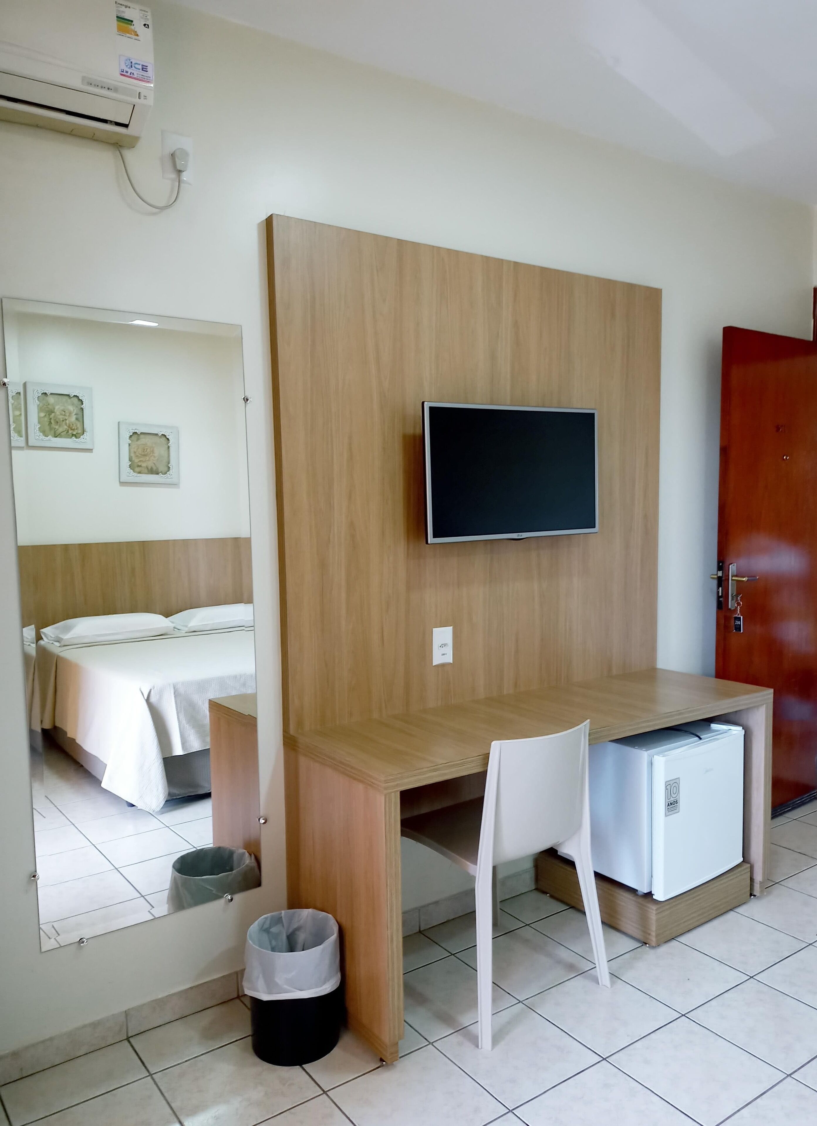 A hotel room with a wooden desk, a television, a small refrigerator, a mirror, and a bed with white bedding.