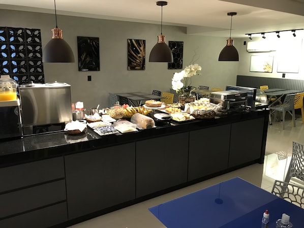A modern kitchen with a buffet setup, featuring a stainless steel refrigerator, a coffee machine, and various food items on the counter.