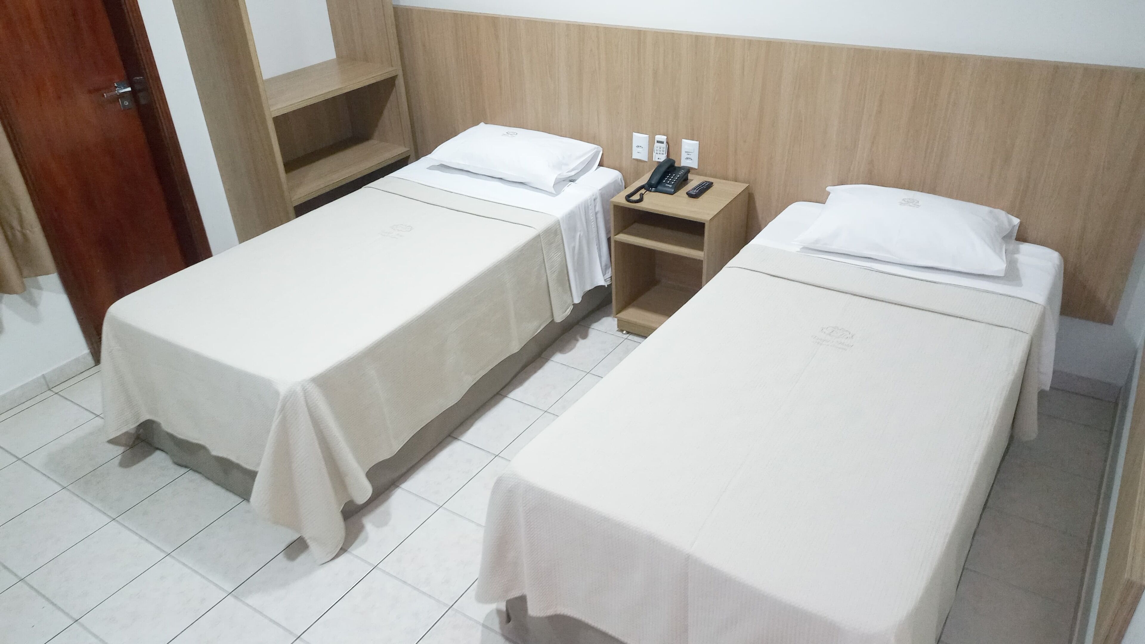 Two single beds with white linens, a wooden headboard, a bedside table with a phone and a remote, and a small shelf.