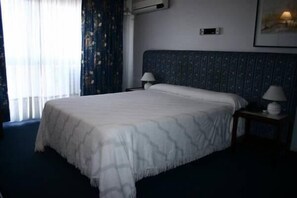 A hotel room with a large bed, two bedside tables with lamps, a window with curtains, and a wall-mounted air conditioning unit.