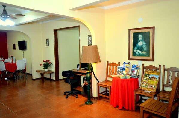 Um saguão de hotel com área de refeições, uma mesa com abajur, uma cadeira com folhetos e um quadro na parede.