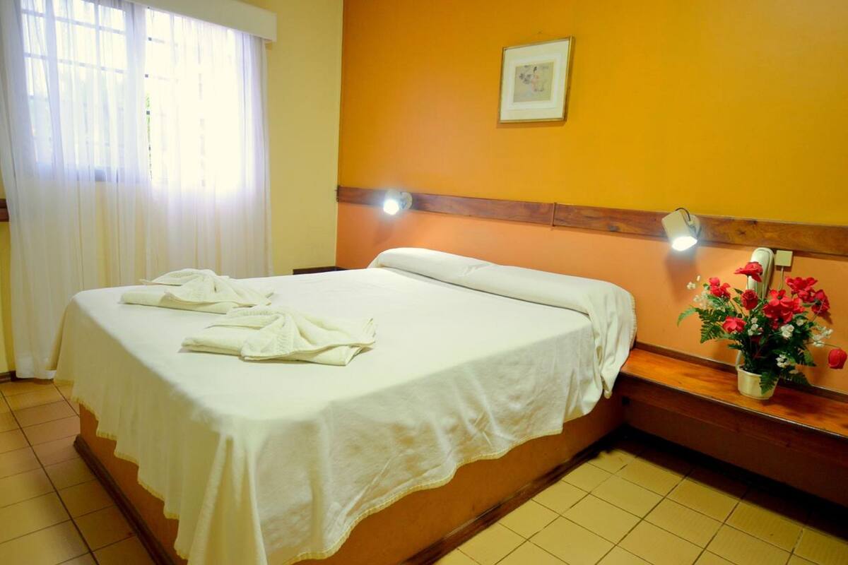 Une chambre d’hôtel avec un lit, une table de chevet avec une lampe, une fenêtre avec des rideaux et un tableau accroché au mur.