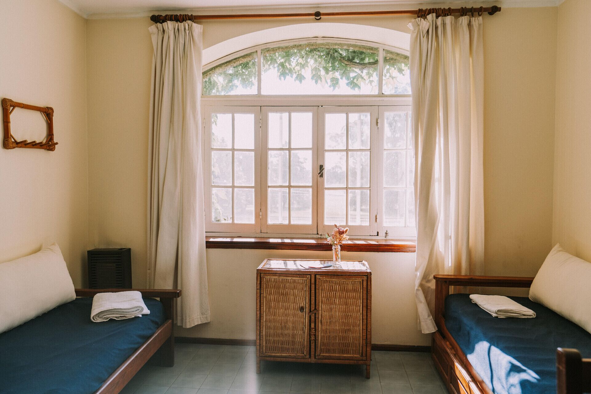 A room with two single beds, a small wicker cabinet, a window with curtains, and a wooden frame on the wall.