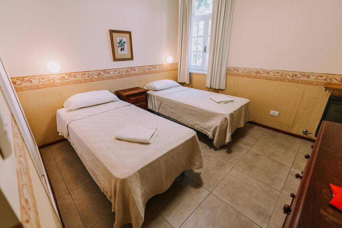 A room with two beds, a small bedside table, a framed picture on the wall, and a window with curtains.