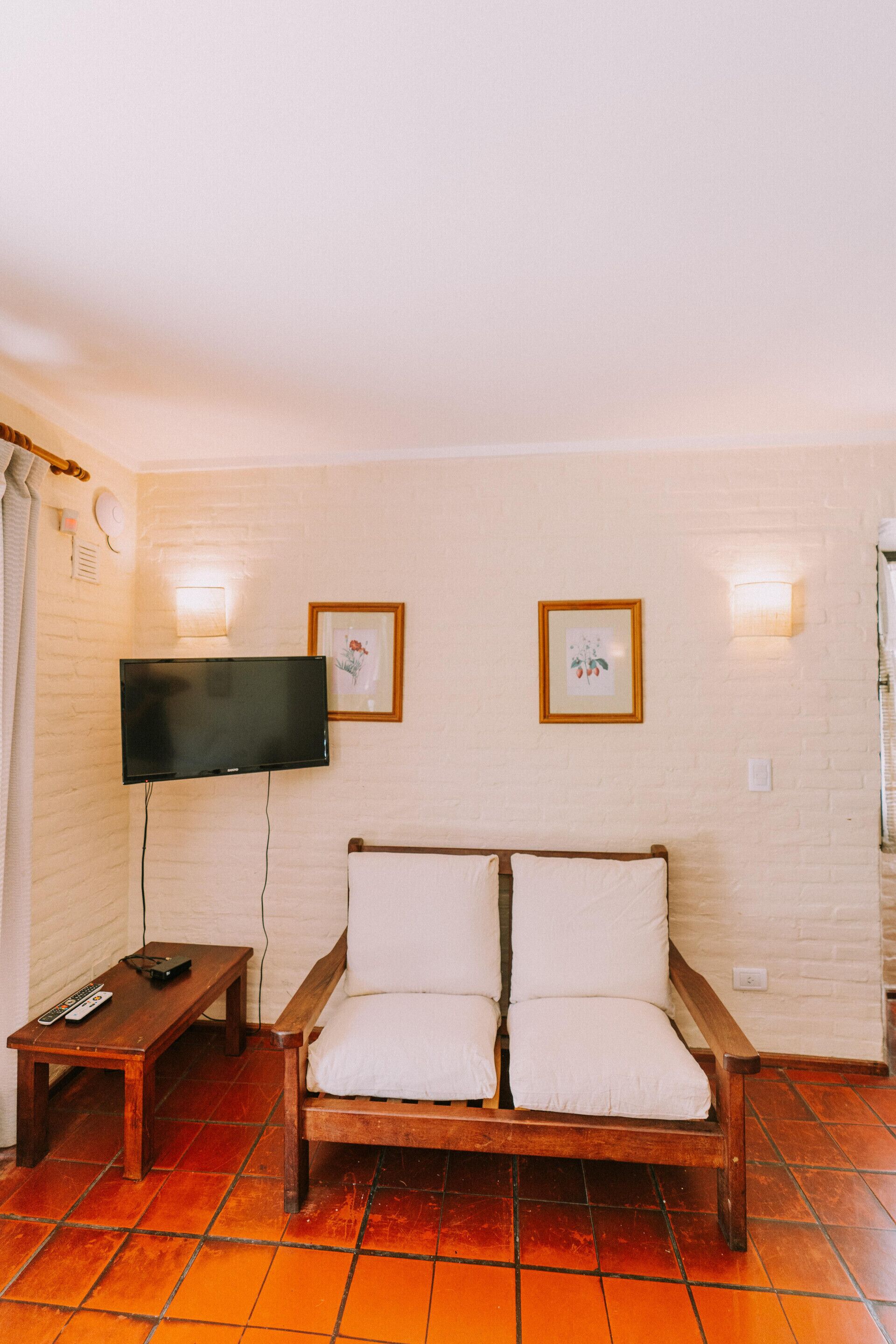 Habitació amb un sofà de fusta, televisió de pantalla plana penjada a la paret, una taula petita de fusta amb un comandament a distància i dos quadres emmarcados a la paret.