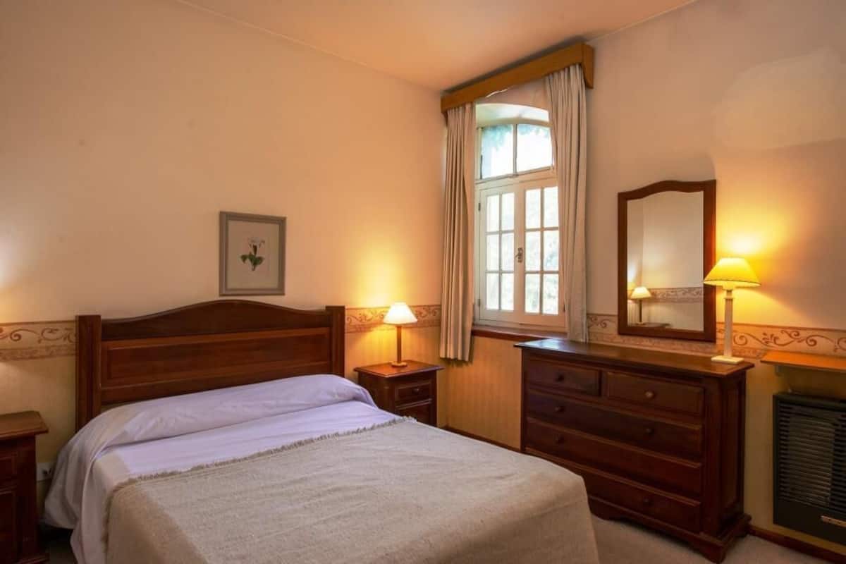 A bedroom with a wooden bed, two bedside tables with lamps, a dresser, a window with curtains, and a framed picture on the wall.