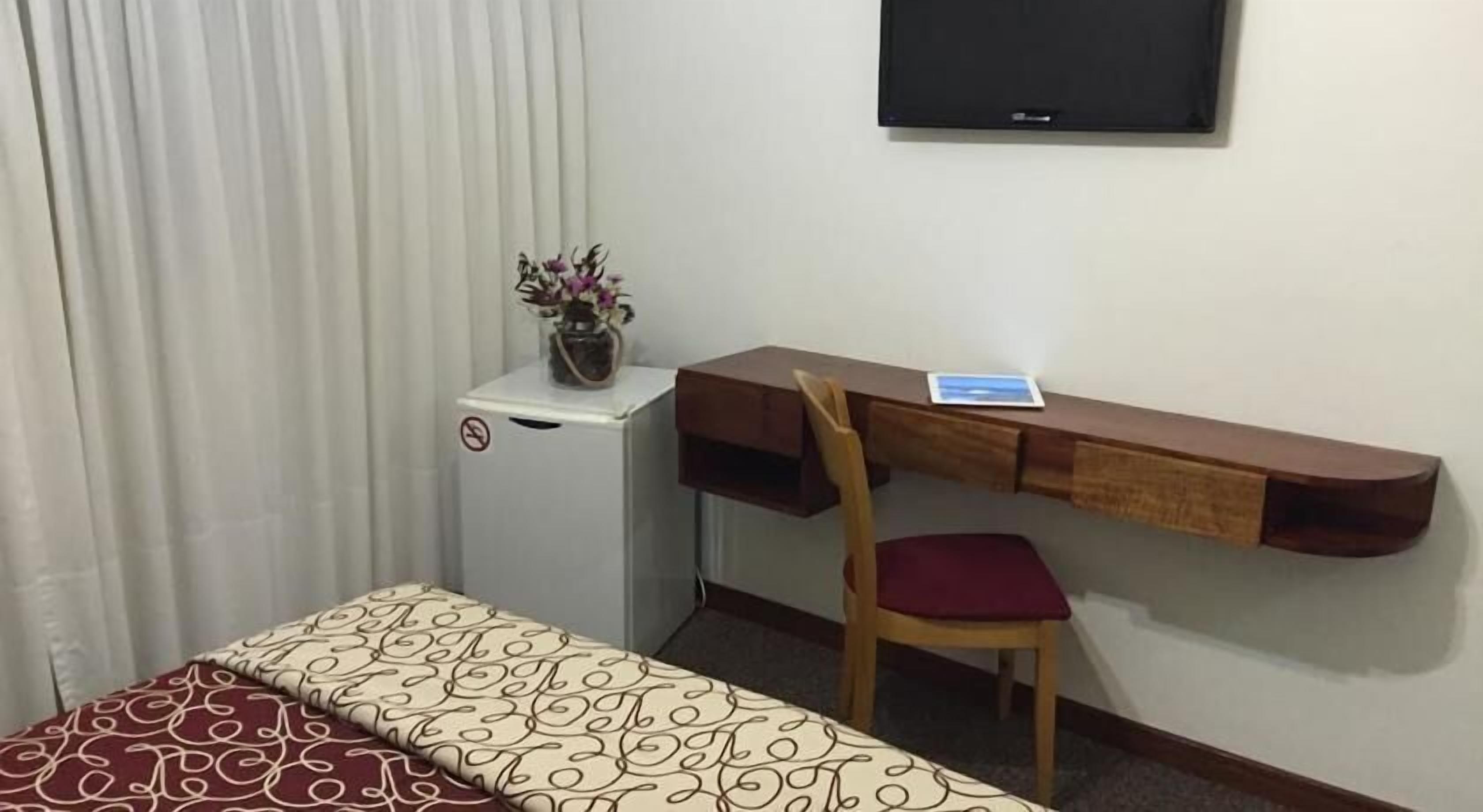Habitación de hotel con cama, escritorio con silla, televisor y refrigerador pequeño.