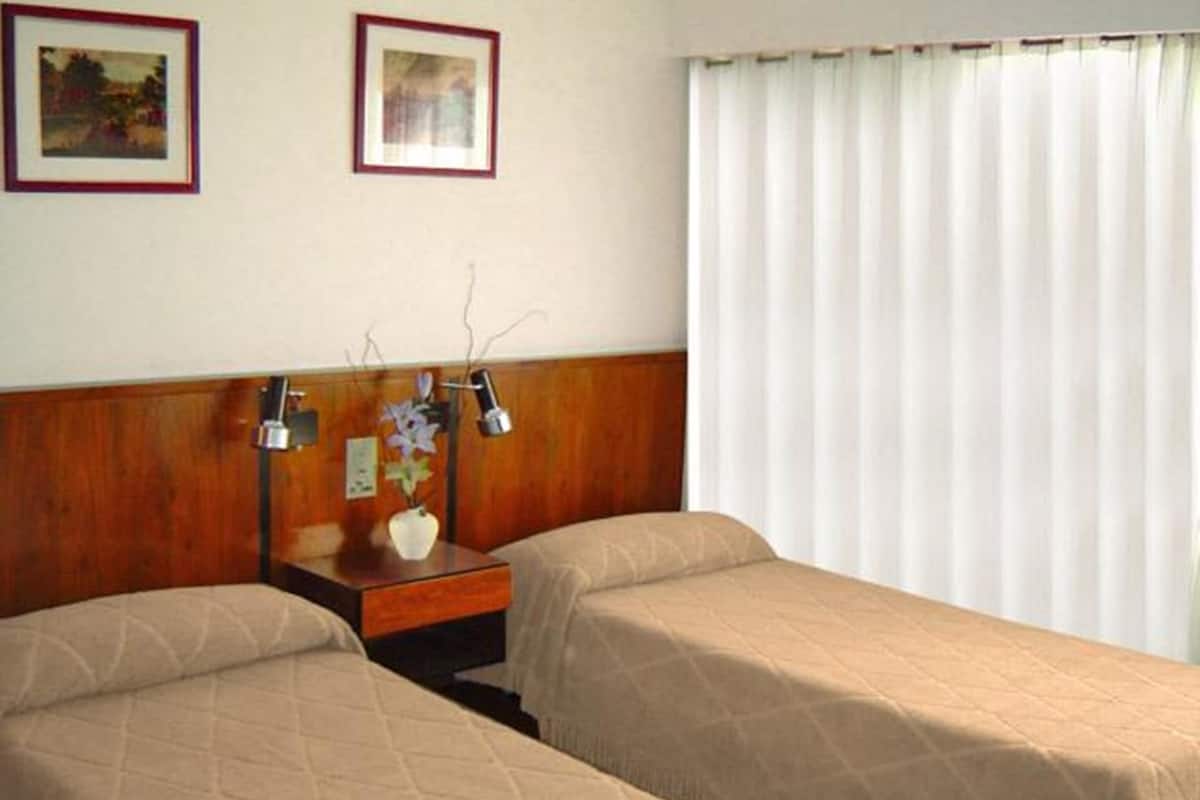 Ein Hotelzimmer mit zwei Betten, einem Holzkopfteil, einem Nachttisch mit Lampe sowie zwei gerahmten Bildern an der Wand.