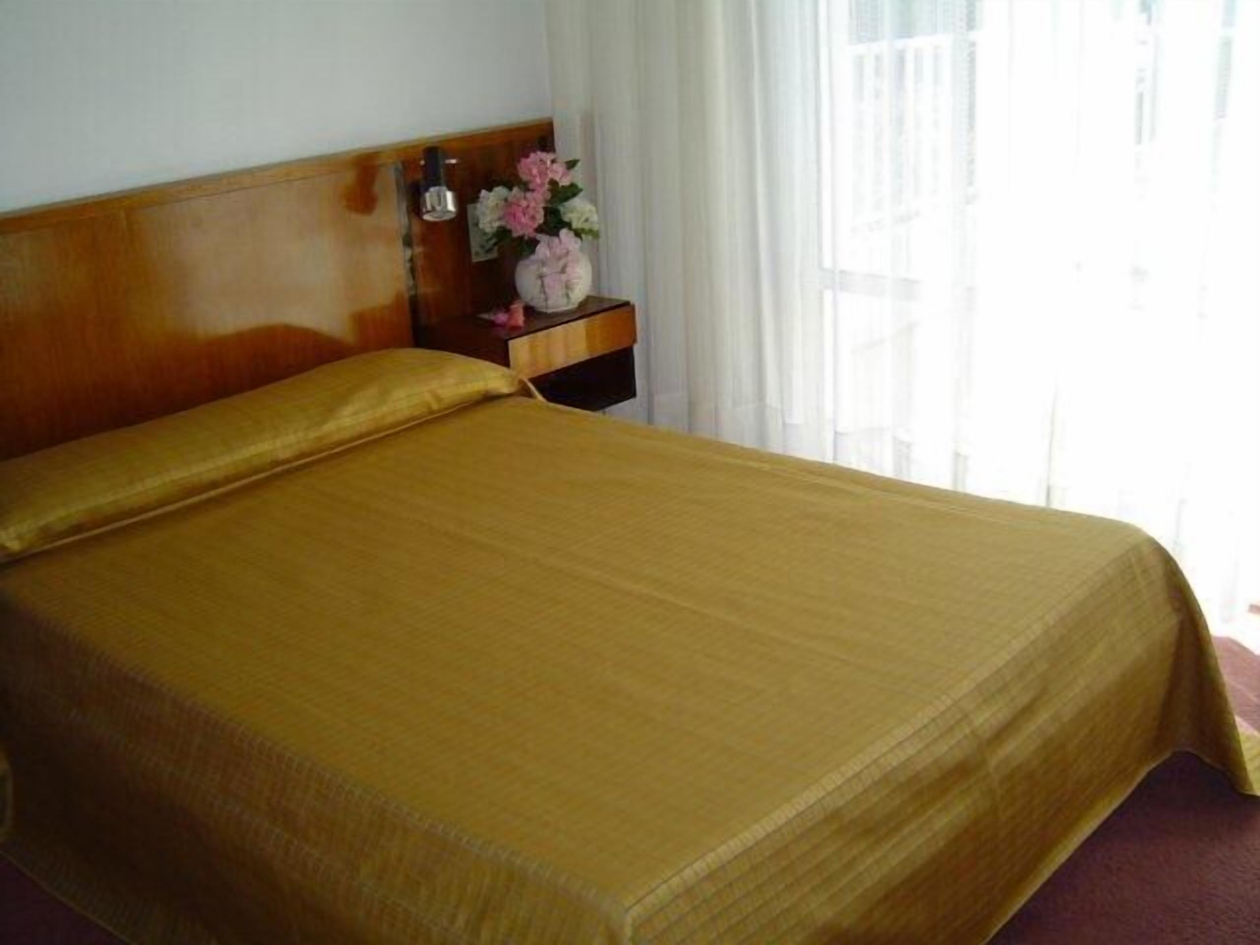 A hotel room with a bed, a nightstand with a vase of flowers, and a window with curtains.