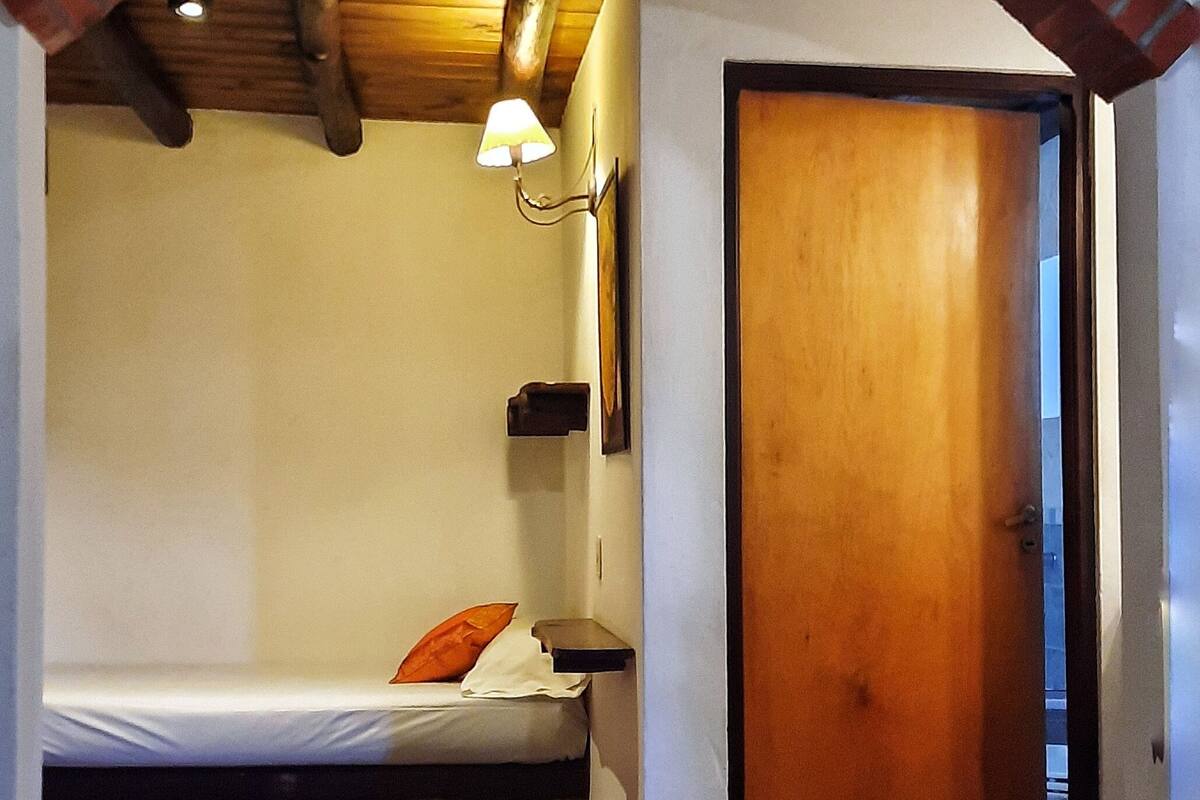 A small, cozy bedroom with a single bed, a wooden door, a wall-mounted shelf, and a ceiling light.