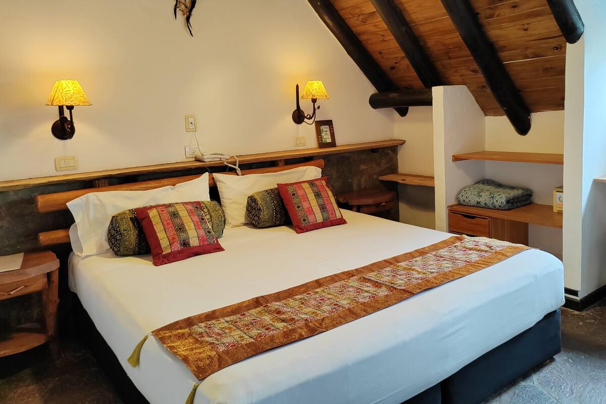A bedroom with a large bed, wooden headboard, two bedside lamps, a wooden nightstand, a small table, and a shelf with a pillow.