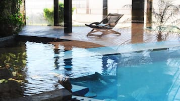 Una piscina interior con terraza de madera, sillas de descanso y plantas en macetas.