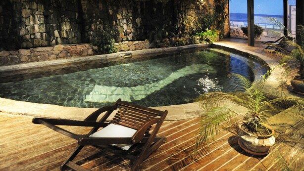 A wooden deck with a pool, stone walls, and a chair.